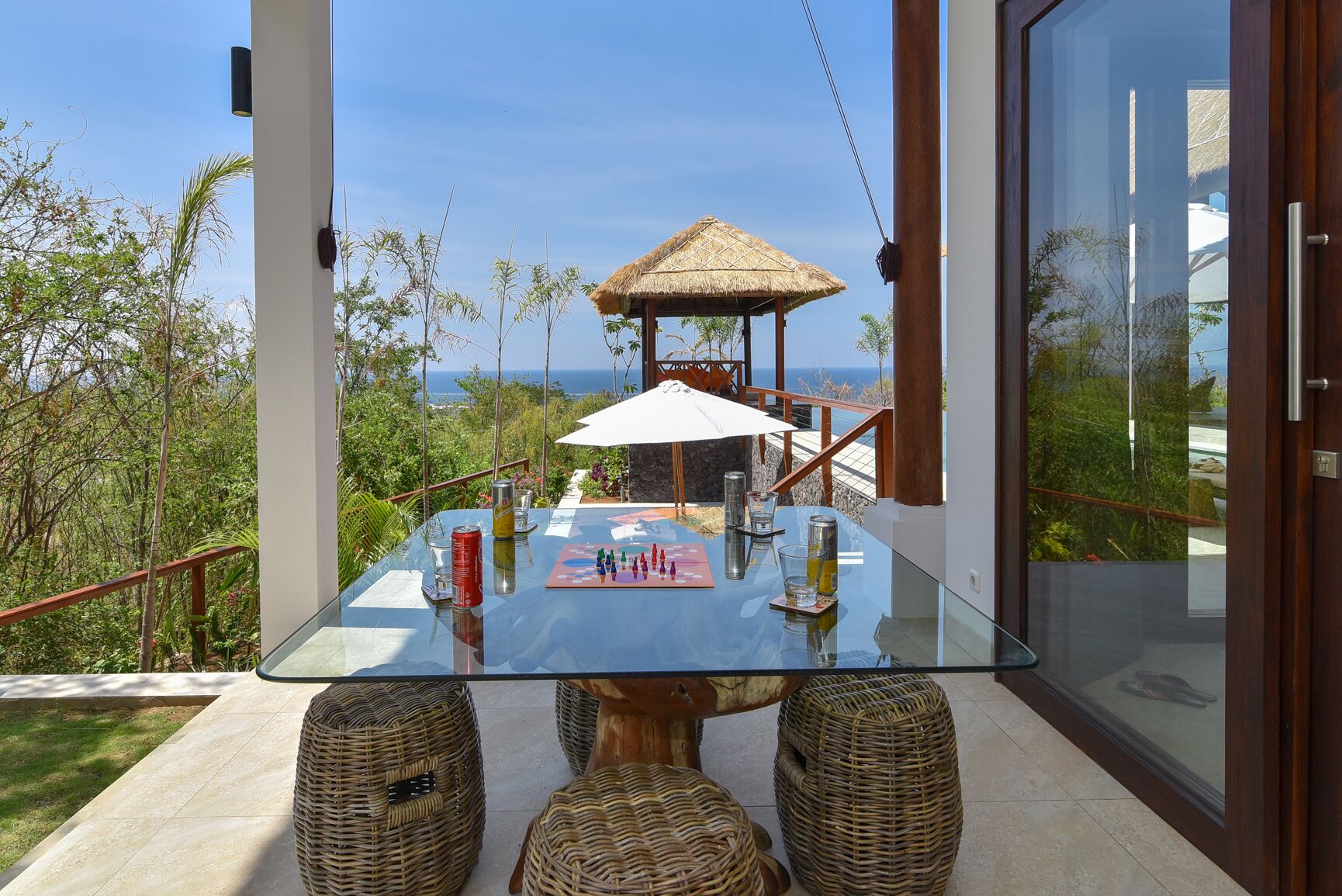 Een glazen tafel met een schaakbord, een gazebo met rieten dak, rieten stoelen en uitzicht op zee.