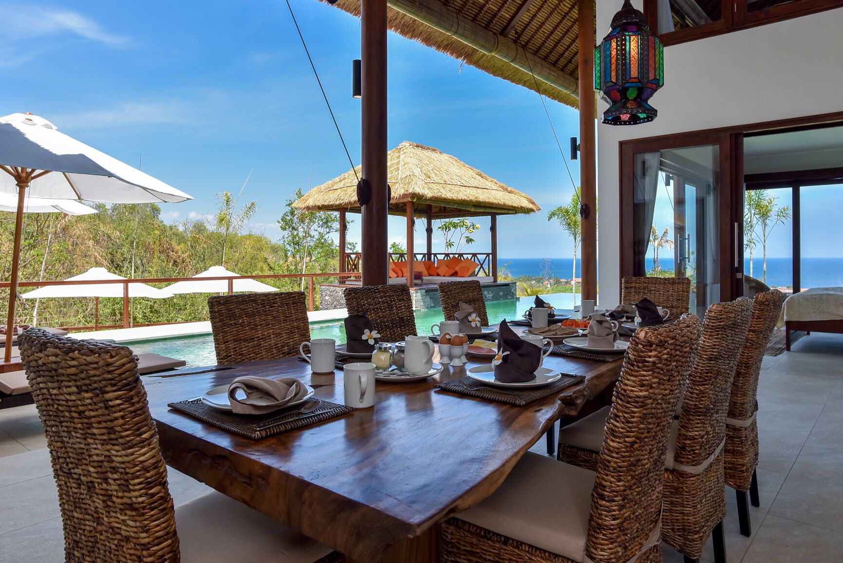 A dining area with a wooden table set for breakfast, overlooking a pool and the ocean.