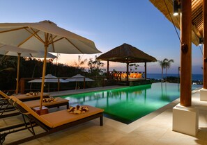 Un espace aménagé au bord de la piscine, avec des chaises longues, des parasols et une structure dotée d’un toit de chaume.