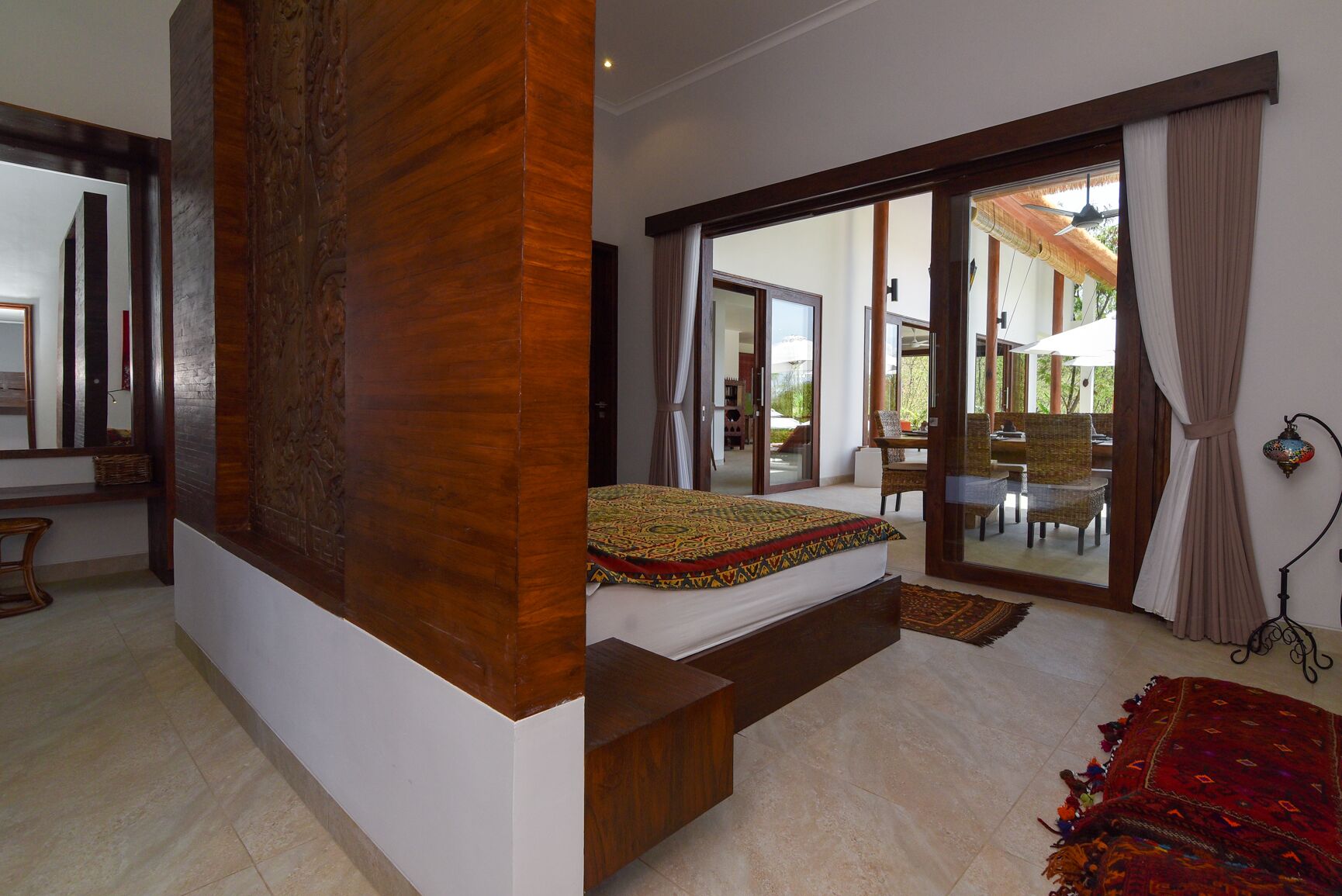 Un dormitorio con una cama grande, un muro decorativo de madera, una puerta corrediza de vidrio que da a un balcón y una alfombra roja estampada.