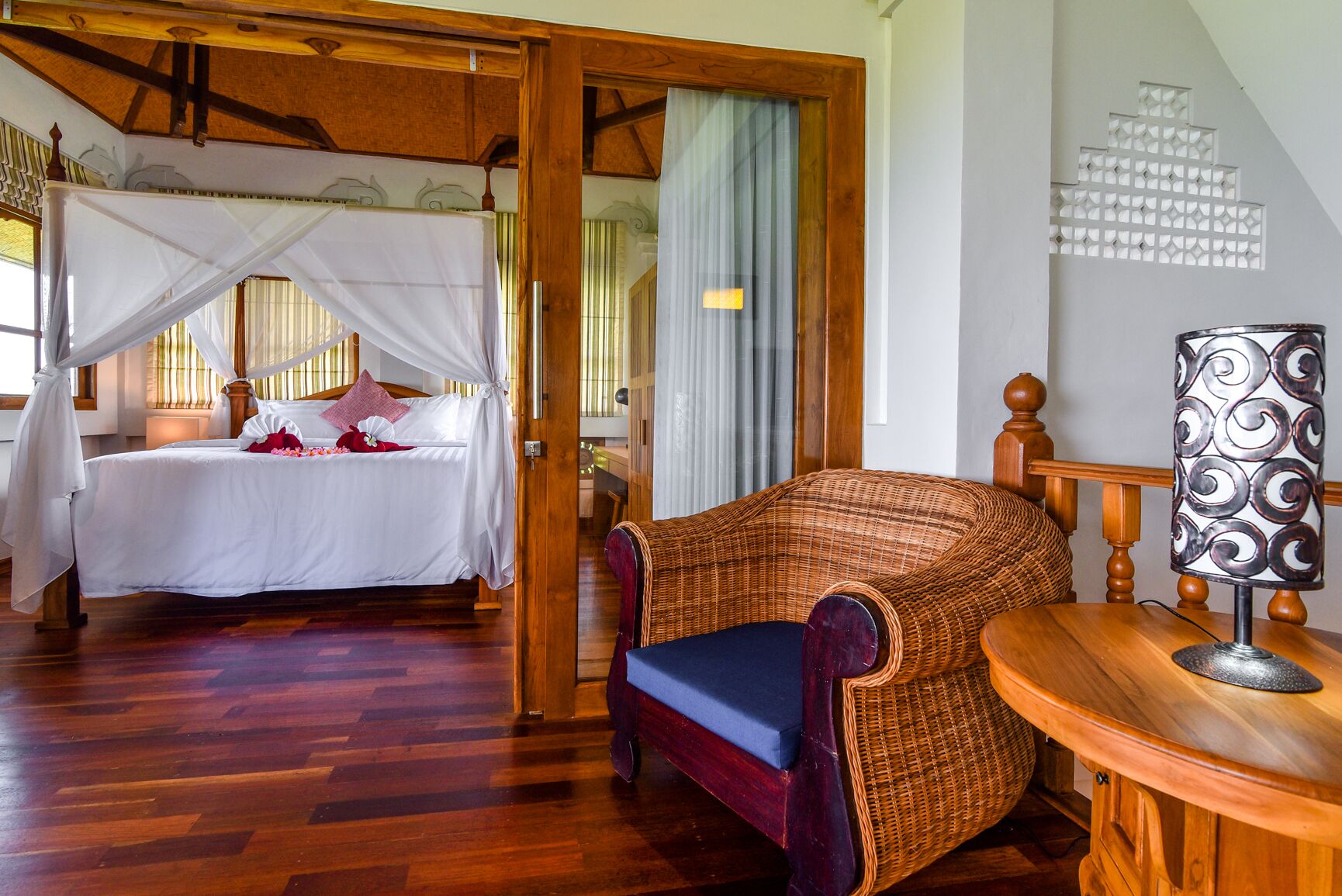 A room with a four-poster bed, a wicker chair, and a table with a lamp.