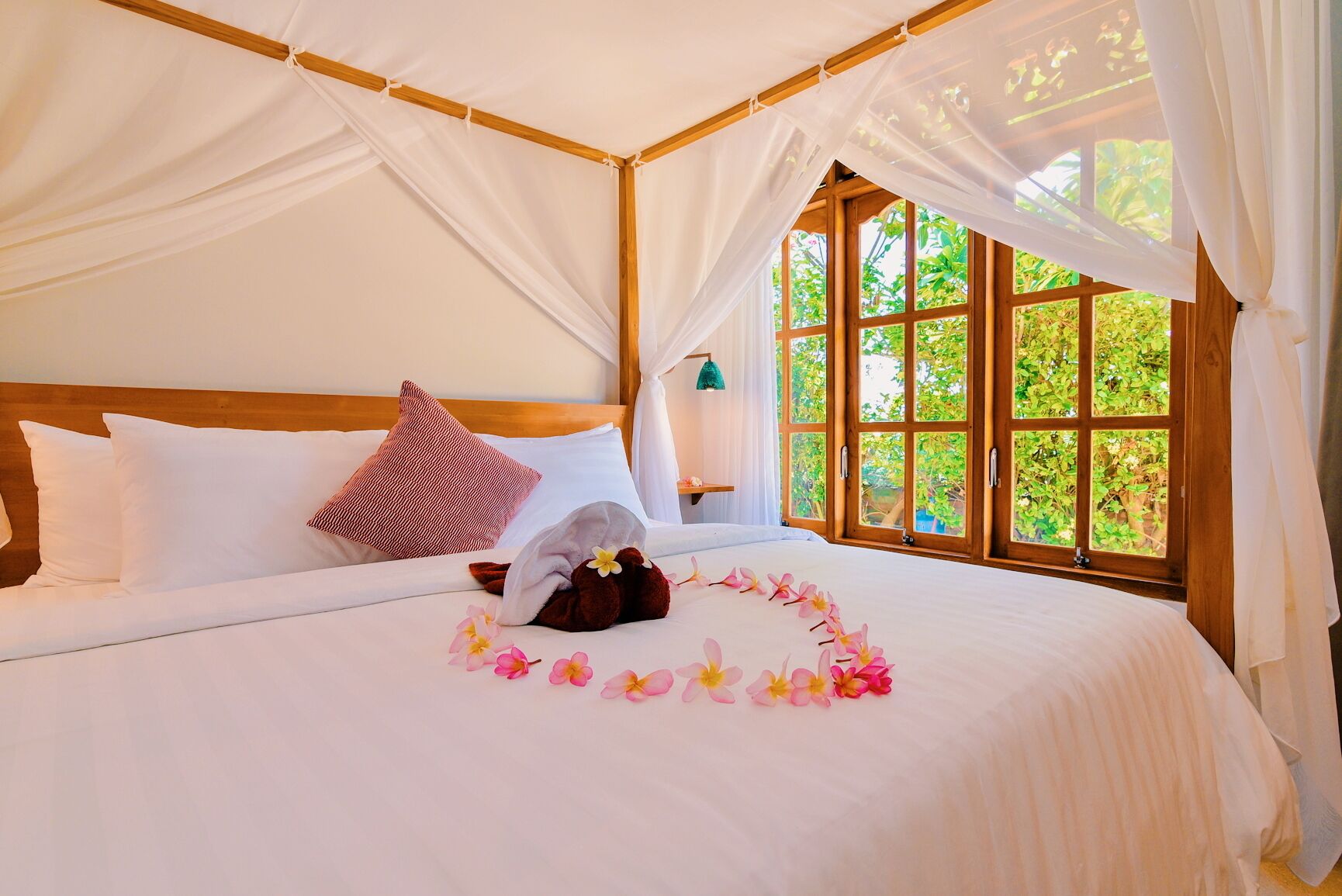 Una cama con sábanas blancas, cabecera de madera, almohada roja y una toalla con flores.