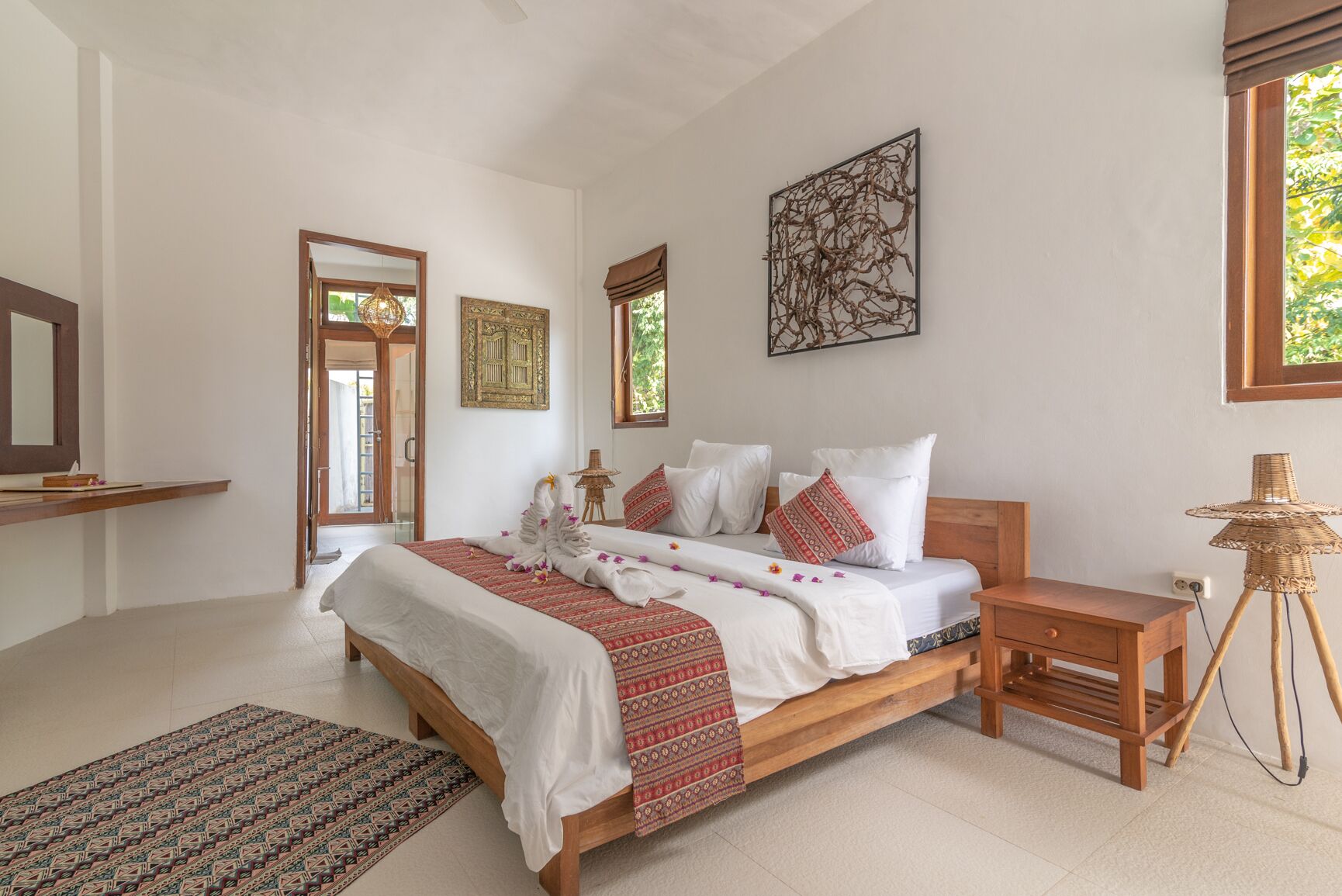 A spacious bedroom with a large bed, a wooden nightstand, a wicker lamp, and a view of greenery outside.