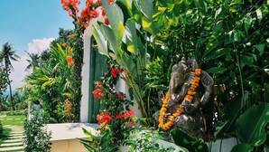 A statue of an elephant deity adorned with garlands, surrounded by lush tropical plants and a pathway leading to a building with green shutters. - Villa Zoubi Bali (Seririt)