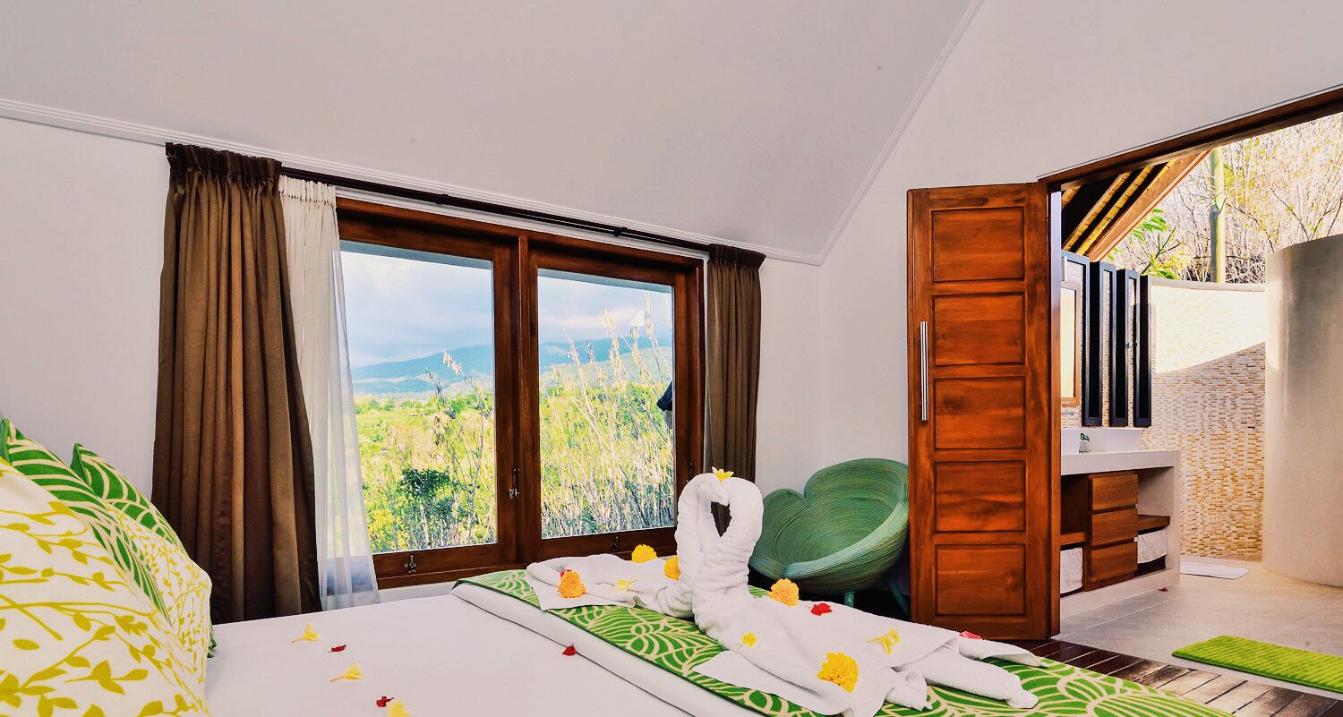 A hotel room with a bed, a green chair, a wooden door, and a view of a landscape through large windows.