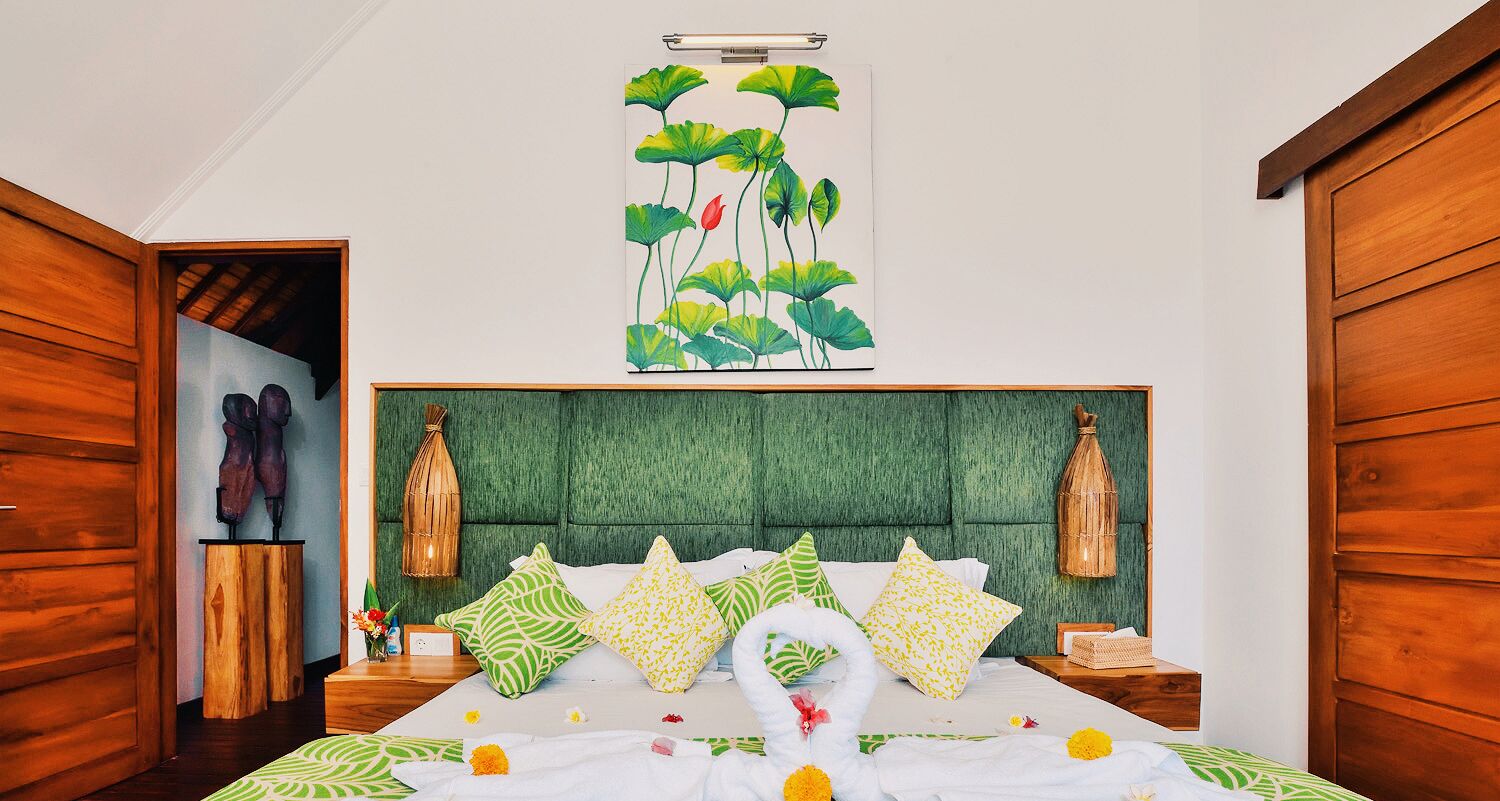 A hotel room with a green upholstered headboard, a bed with white bedding and patterned pillows, a wall-mounted artwork of lotus flowers, and wooden doors.