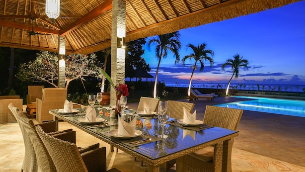 An outdoor dining area with wicker furniture and a glass table, overlooking a pool and palm trees at dusk. - Villa Duyung (Seririt)