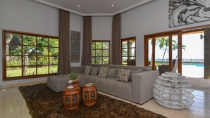 A spacious living room with a grey sofa, a large window with a view of greenery, and a decorative white metal sculpture. - Villa Duyung (Seririt)