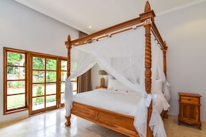 A four-poster bed with a canopy, a wooden nightstand, and a view of a garden through a large window. - Villa Duyung (Seririt)