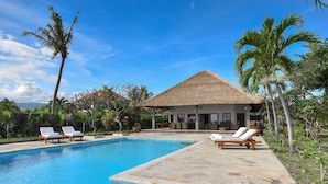 A poolside view of a resort with a thatched-roof building, lounge chairs, and tropical vegetation. - Villa Duyung (Seririt)