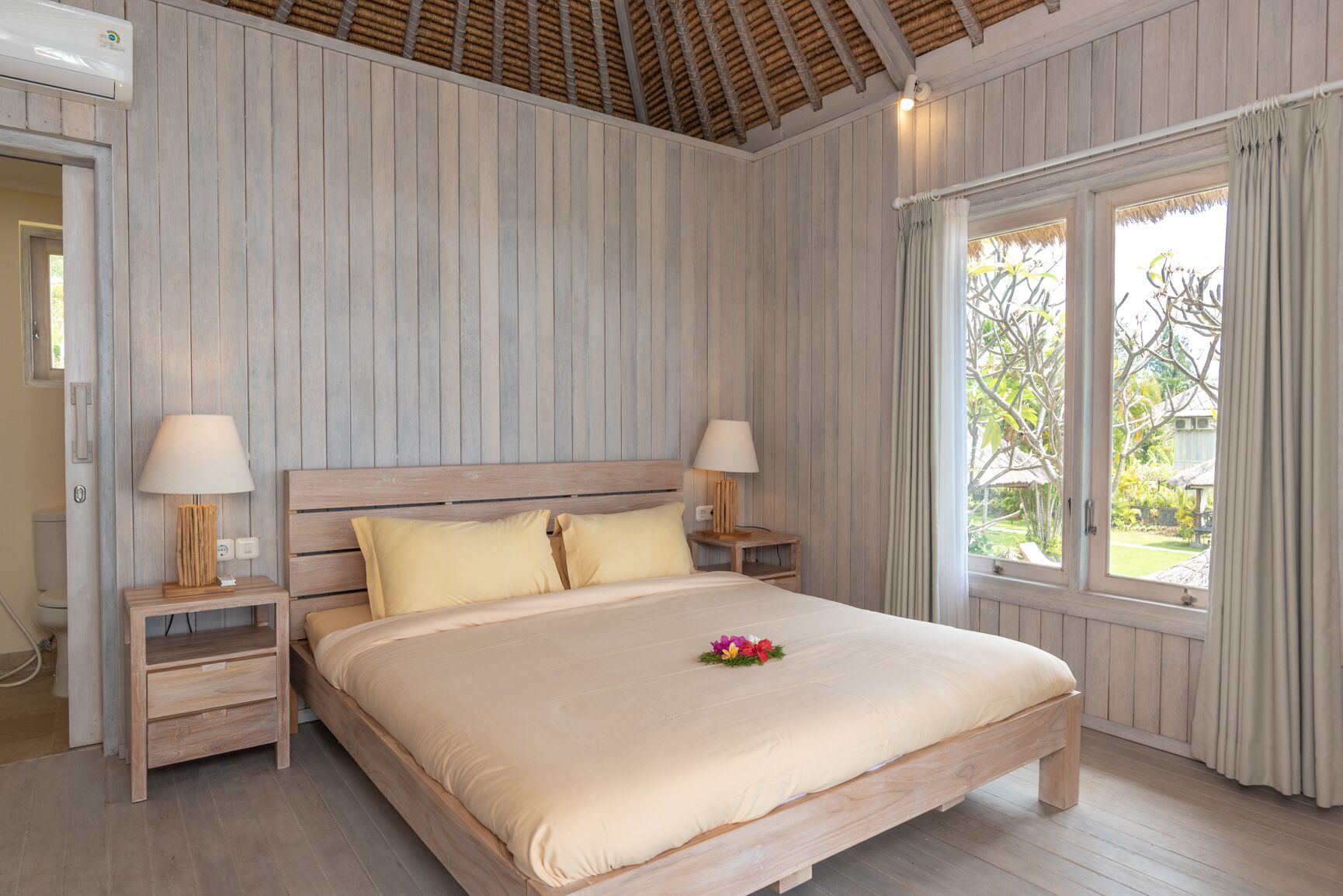 A bedroom with a wooden bed, two bedside tables with lamps, a window with curtains, and a view of greenery outside.