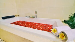 A bathtub filled with red pebbles, a black bath mat, a white towel with yellow flowers, and a small plant beside it. - Villa Banyan (Seririt)