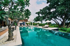A swimming pool with lounge chairs and umbrellas, surrounded by trees and a modern building.