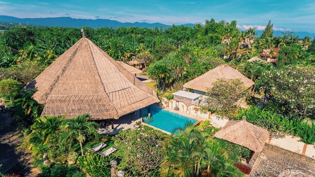 Ein Resort mit einem großen, mit Schilfdächern versehenen Gebäude, einem Swimmingpool und üppiger tropischer Vegetation.