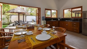 Un coin repas avec une table dressée pour quatre personnes, une cuisine dotée de meubles en bois et une vue sur un jardin agrémenté d’une structure à toit de chaume.