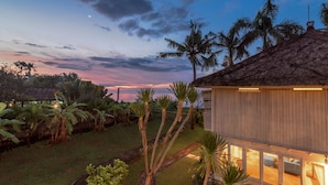 A house with a thatched roof, surrounded by a well-maintained garden with palm trees and a view of the ocean at dusk. - Villa Lumbung (Sukasada)