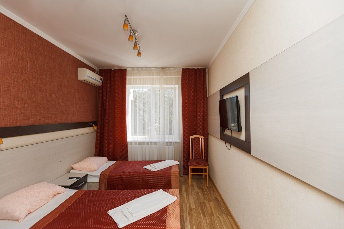 A hotel room with two beds, a TV mounted on the wall, a chair, and a window with curtains.