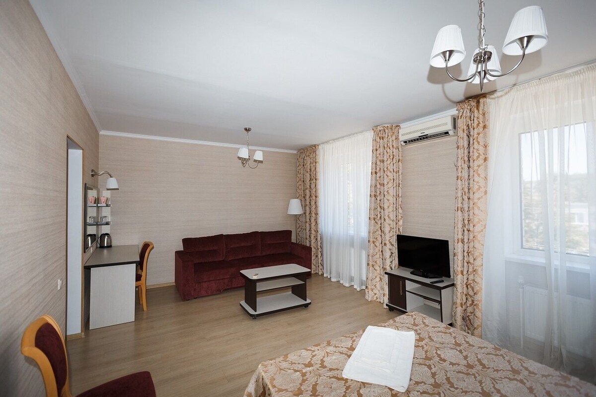 A hotel room with a sofa, TV, dining table, and a small desk with a mirror and lamp.