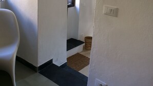 Un pasillo con pared blanca, una silla blanca, una ventana pequeña y una puerta.