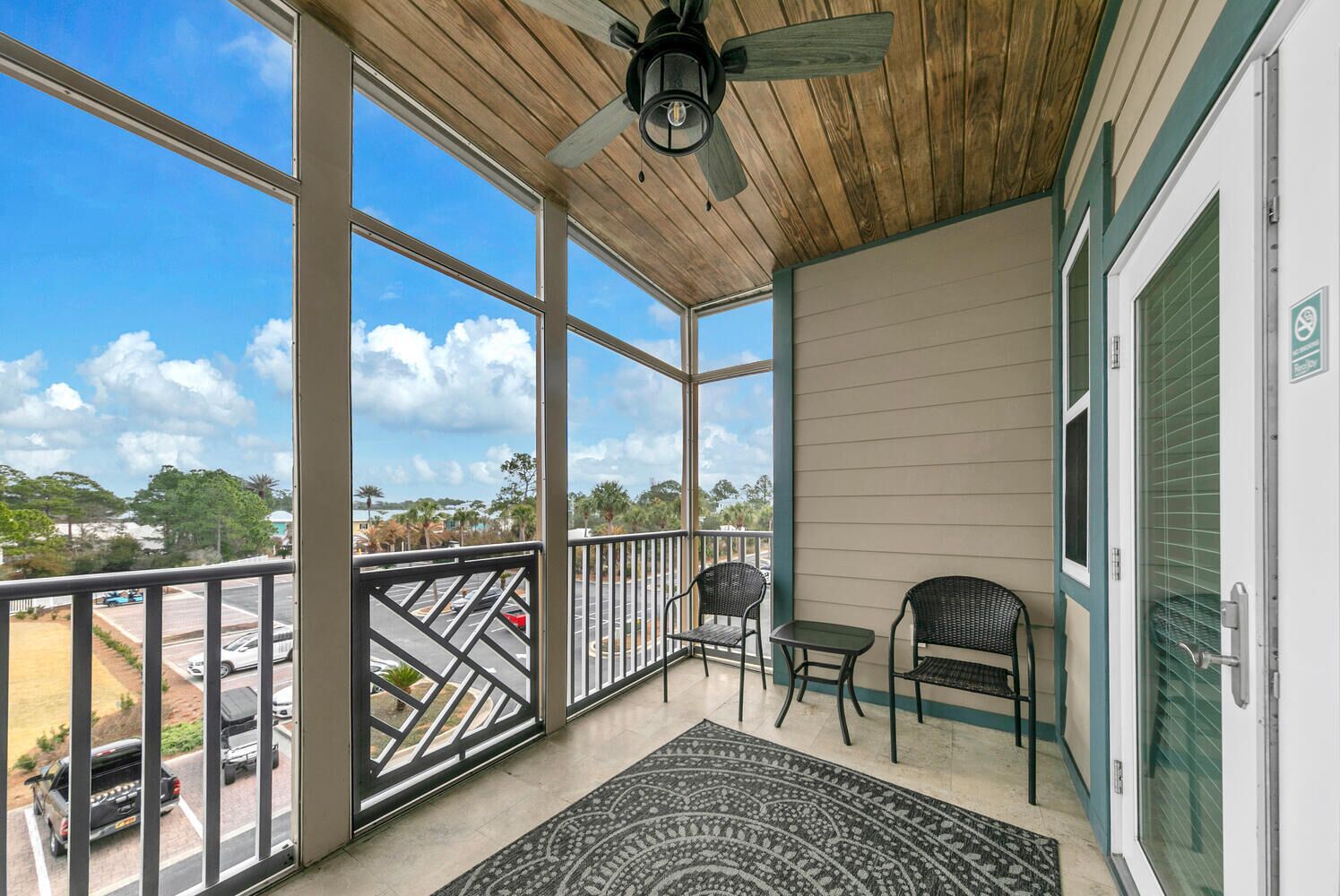 A covered balcony with a ceiling fan, two chairs, a small table, and a view of a parking lot and trees.