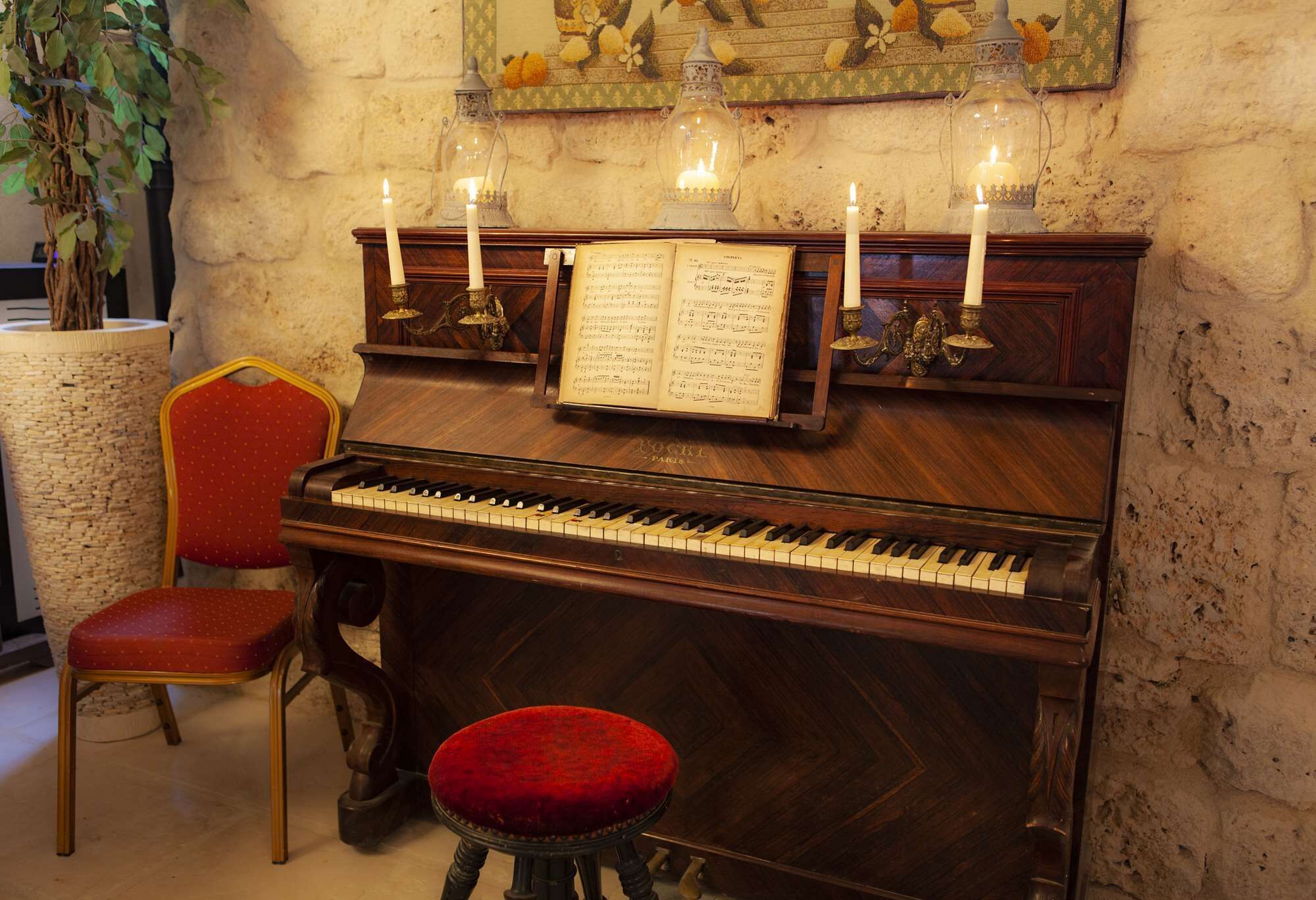 A grand piano with sheet music, candles, and a red upholstered stool in a room with stone walls and a plant.