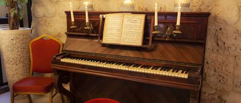 A grand piano with sheet music, candles, and a red upholstered stool in a room with stone walls and a plant.