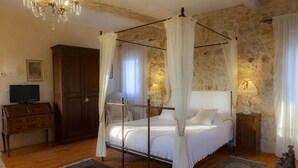 A bedroom with a four-poster bed, a wooden dresser, a television, and a stone wall.