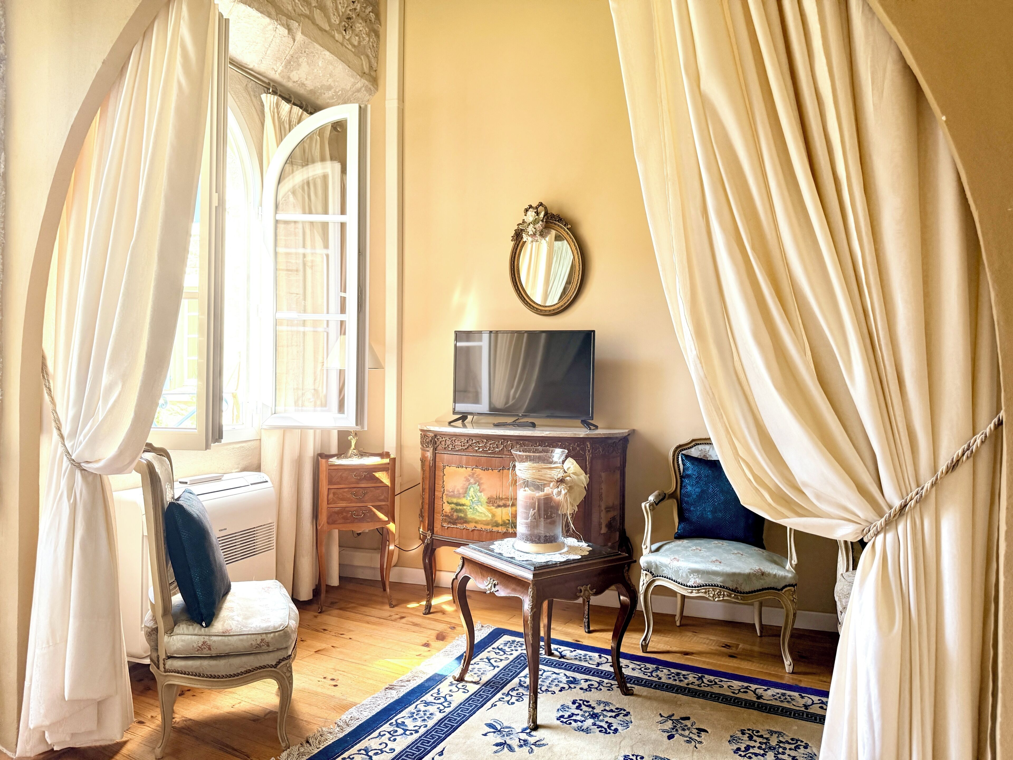 Una habitación con una televisión sobre un mueble de madera, una silla con cojín, una mesita con un florero y una alfombra estampada.