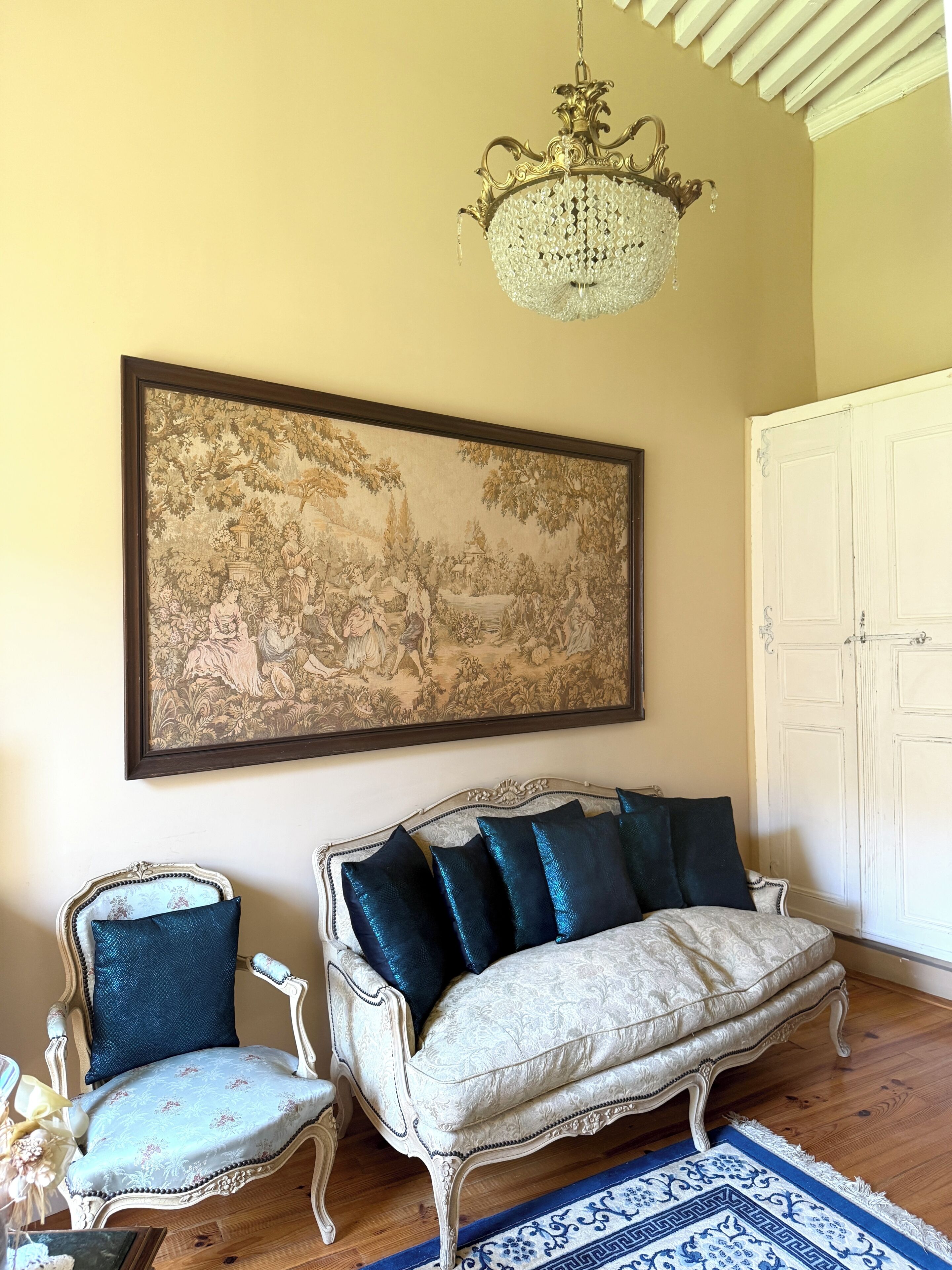 Una habitación con un cuadro de paisaje grande enmarco, un sofá blanco con cojines oscuros, una silla antigua y una araña.