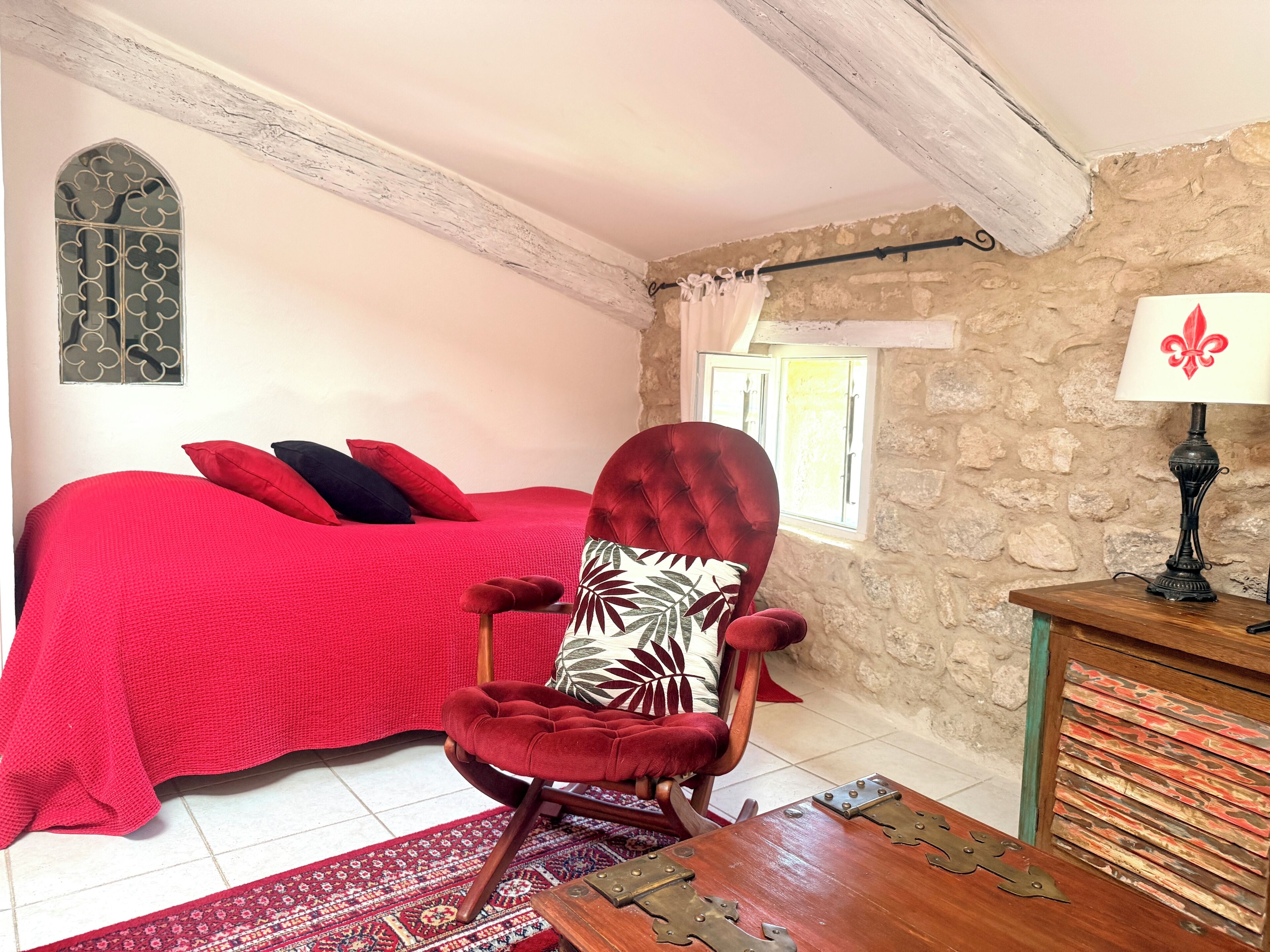 A bedroom with a red bedspread, a red chair with a patterned cushion, a wooden nightstand, a stone wall, and a window with curtains.