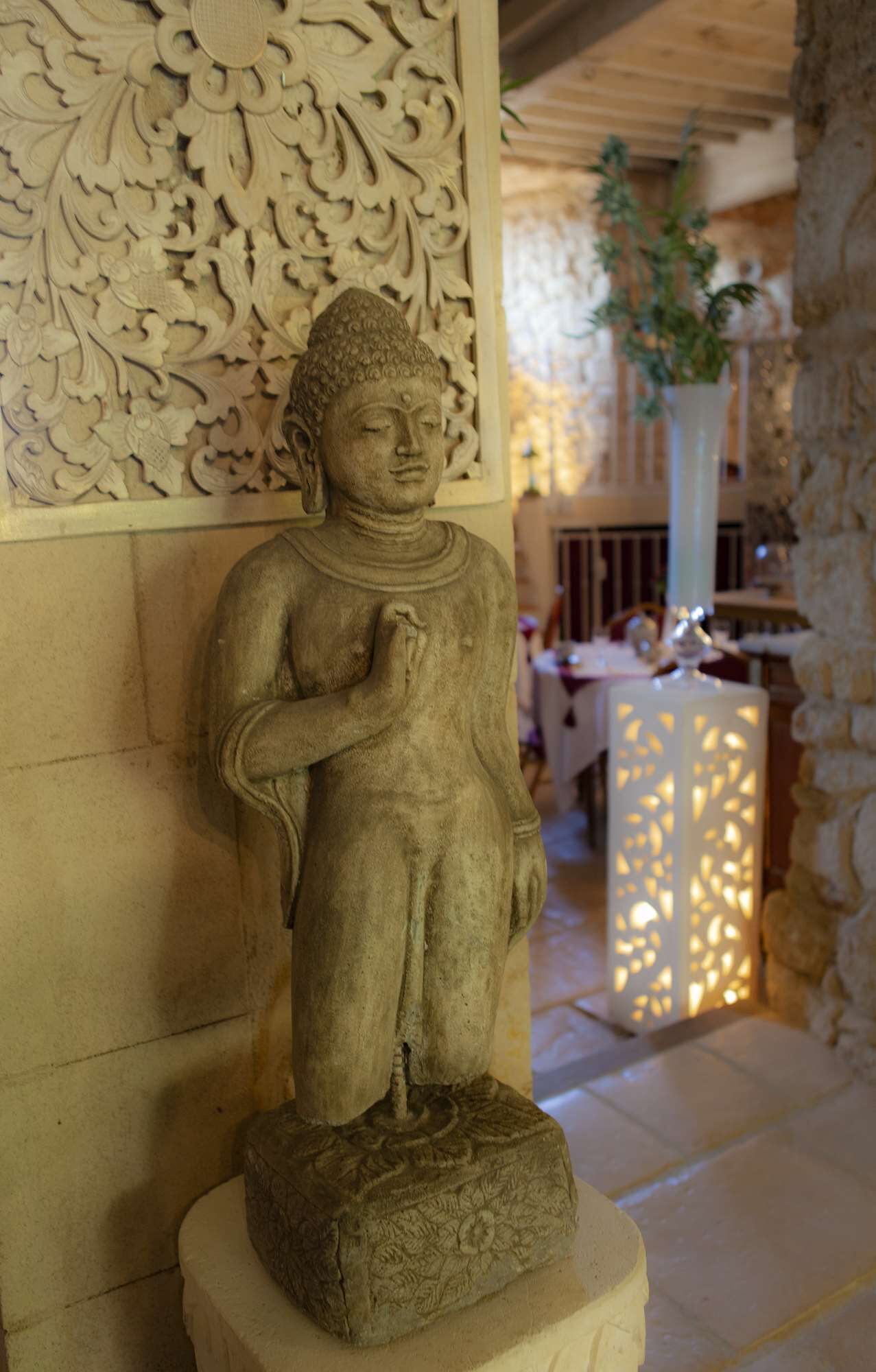 Une statue de Bouddha en pierre, dans une pièce ornée de sculptures complexes sur les murs et agrémentée d’une plante en pot.