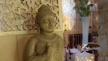 Une statue de Bouddha en pierre, dans une pièce ornée de sculptures complexes sur les murs et agrémentée d’une plante en pot.