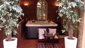 A spa area with a hot tub, two chairs, a table with a tray, and two large potted plants.