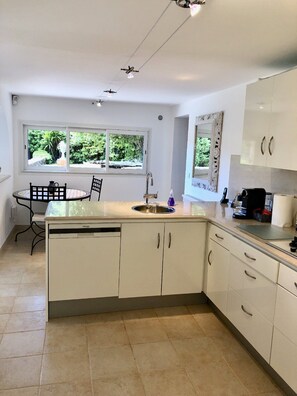 A modern kitchen with white cabinetry, a central island, a dining area with a table and chairs, and a view of greenery through the windows.