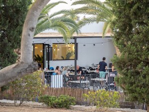 An outdoor dining area with tables and chairs, surrounded by a wooden fence and palm trees. - --- Wood Mobile Home 1 (Calig/Castellon)