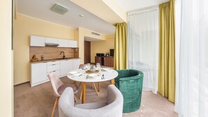 Een moderne hotelkamer met een eethoek, een groene stoel, een witte tafel gedekt voor vier personen en een groot raam met gele gordijnen.