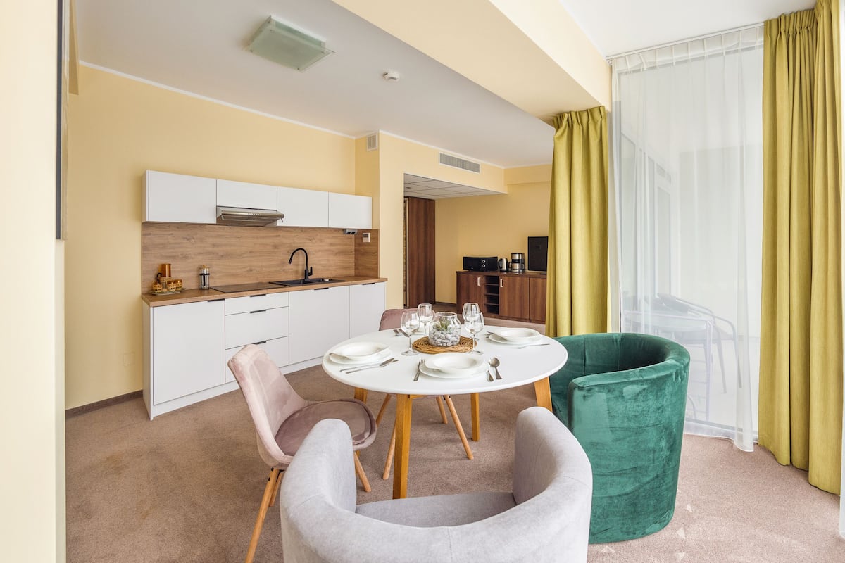 Une chambre d’hôtel moderne avec un coin repas, un fauteuil vert, une table blanche dressée pour quatre personnes et une grande fenêtre ornée de rideaux jaunes.