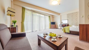 Hotelkamer met een bank, een houten tafel met kopjes en citroenen, een bed met een groene deken, een televisie, een bureau en een groot raam met gordijnen.