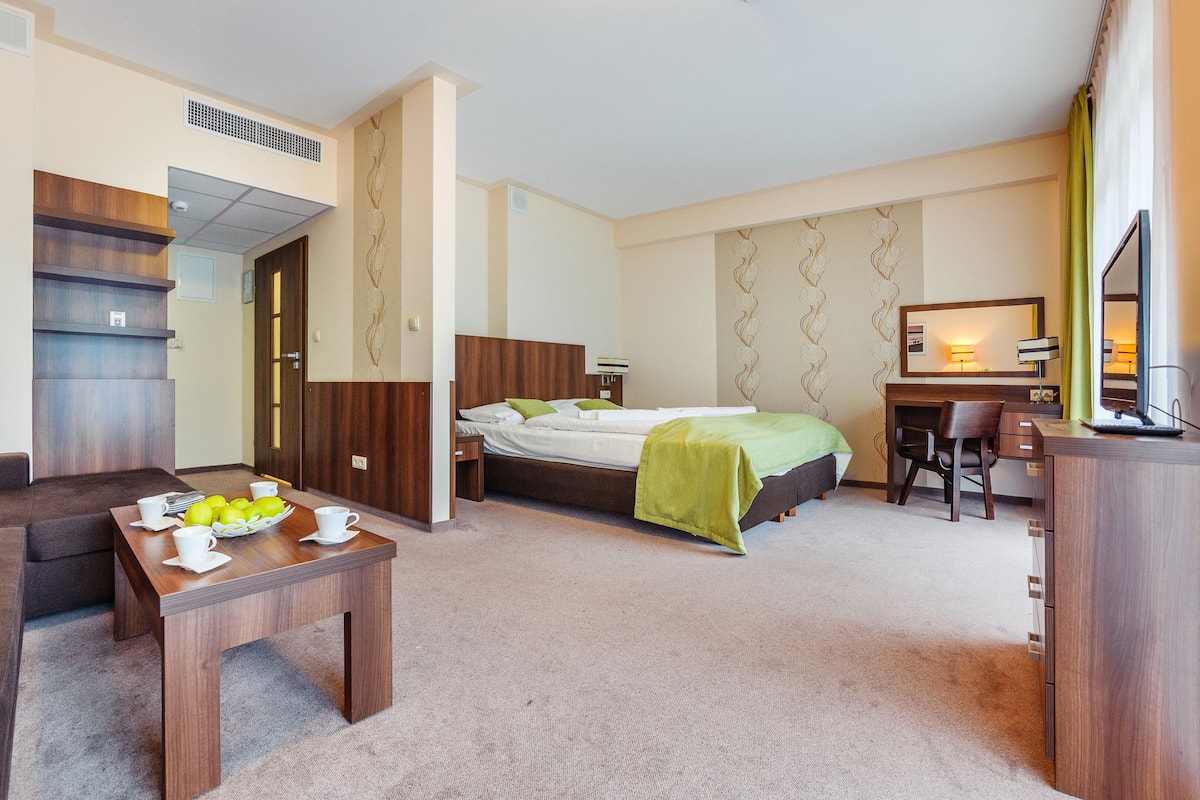 Une chambre d’hôtel avec un lit, un bureau, une chaise, un canapé et une petite table sur laquelle se trouvent des tasses et des pommes.