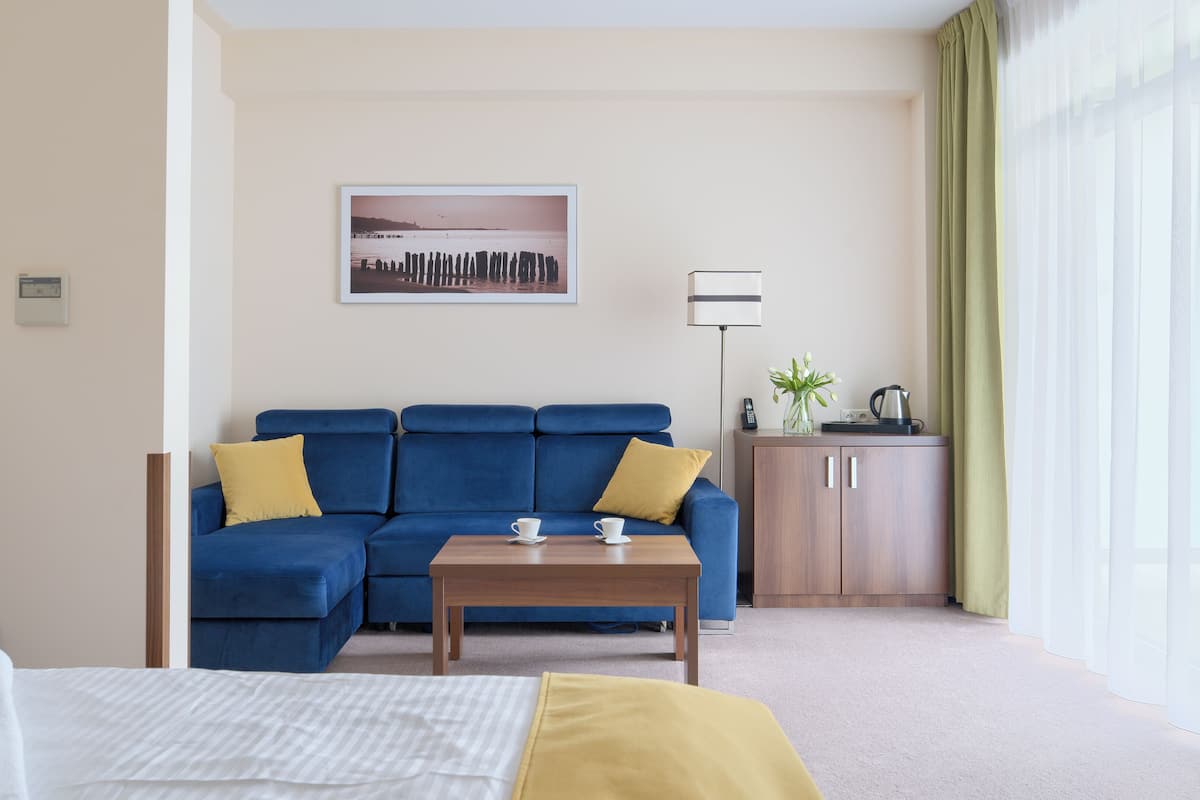 Une chambre d’hôtel avec un canapé bleu, une table basse en bois, un lit, une armoire et une fenêtre ornée de rideaux légers.