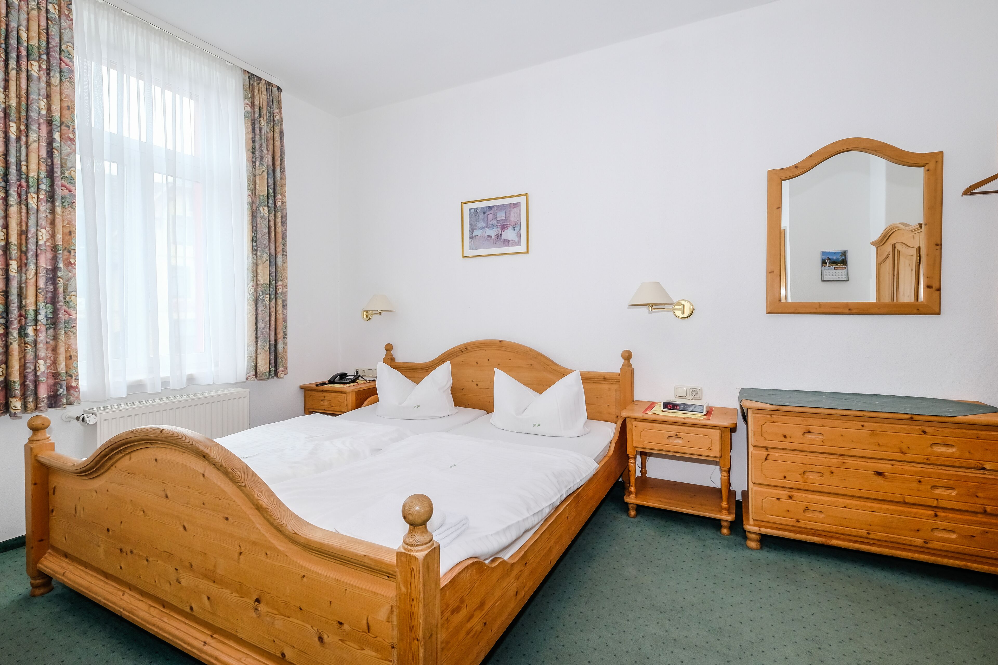 A wooden bed with white bedding, a wooden dresser, a nightstand, a mirror, and a window with curtains.