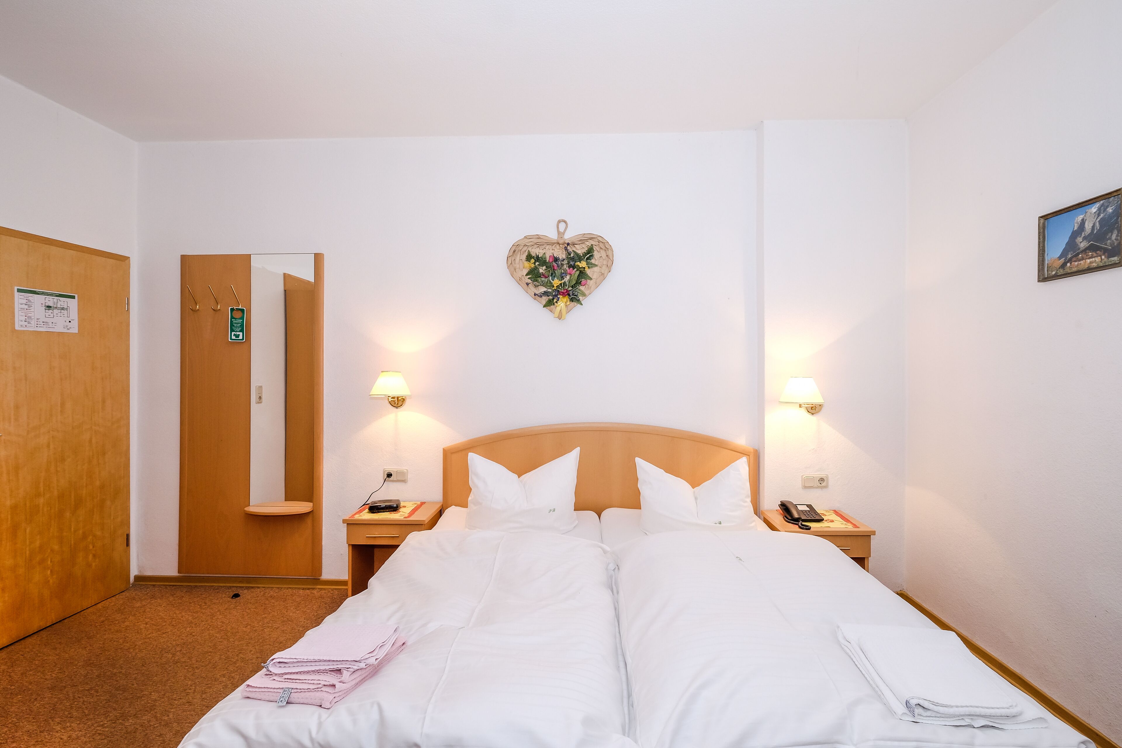 Quarto de hotel com uma cama grande, duas mesas de cabeceira com abajures, uma porta de madeira, um espelho e uma decoração em formato de coração na parede.