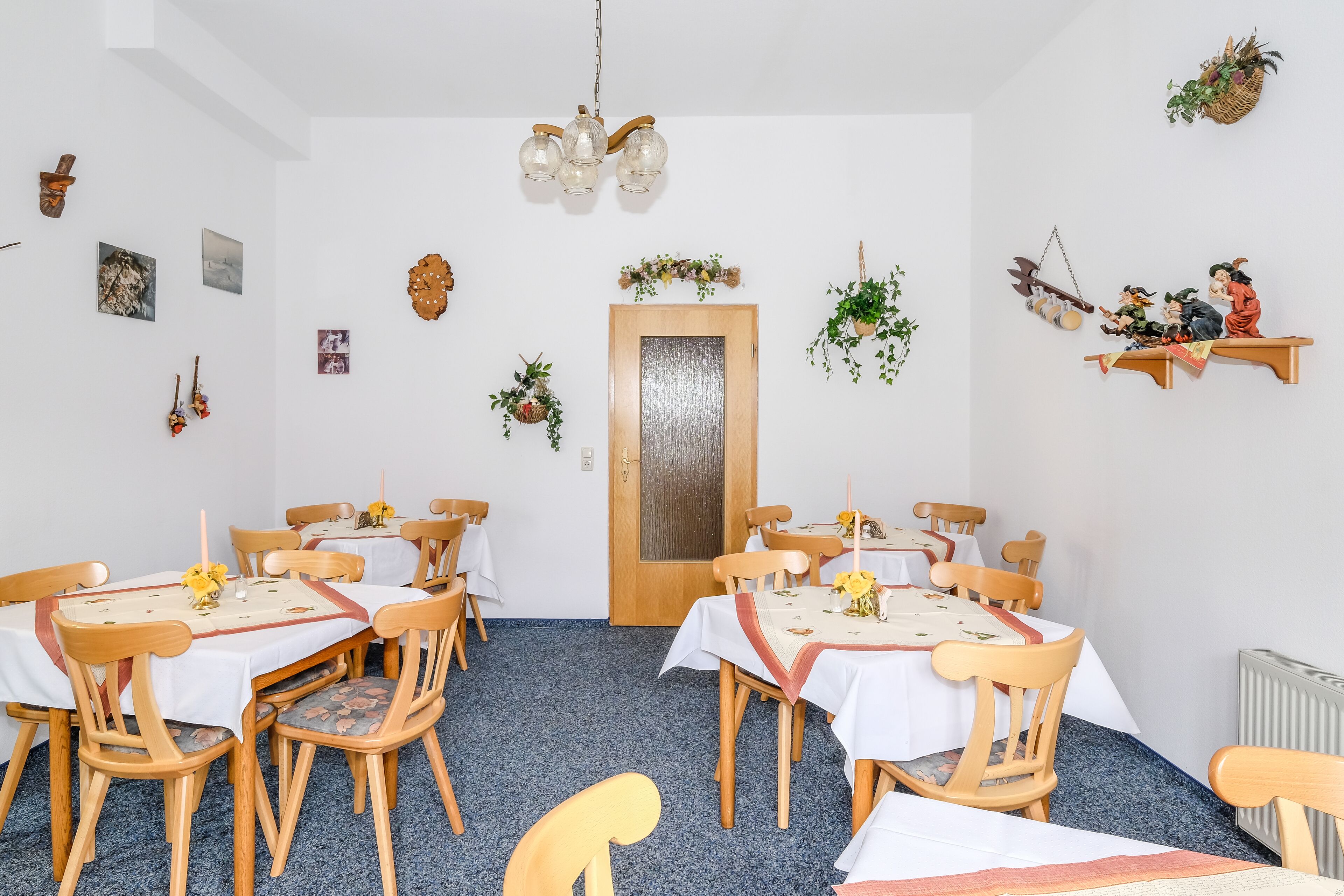 A dining area with wooden chairs and tables, a chandelier, and wall decorations including pictures and plants.