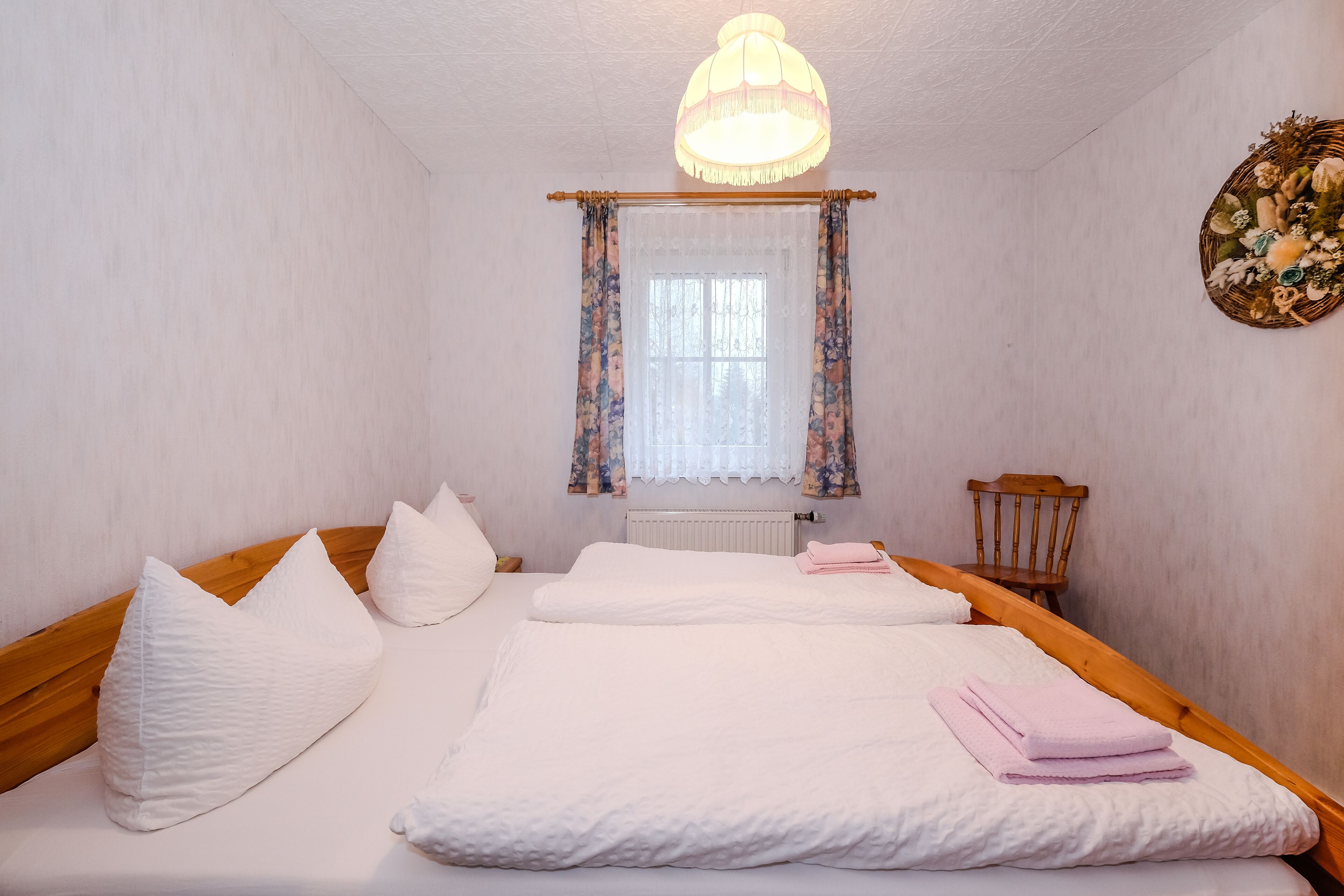 Una cama bien hecha con sábanas y almohadas blancas, un cabecero de madera, una silla de madera, una ventana con cortinas florales y una lámpara colgante.