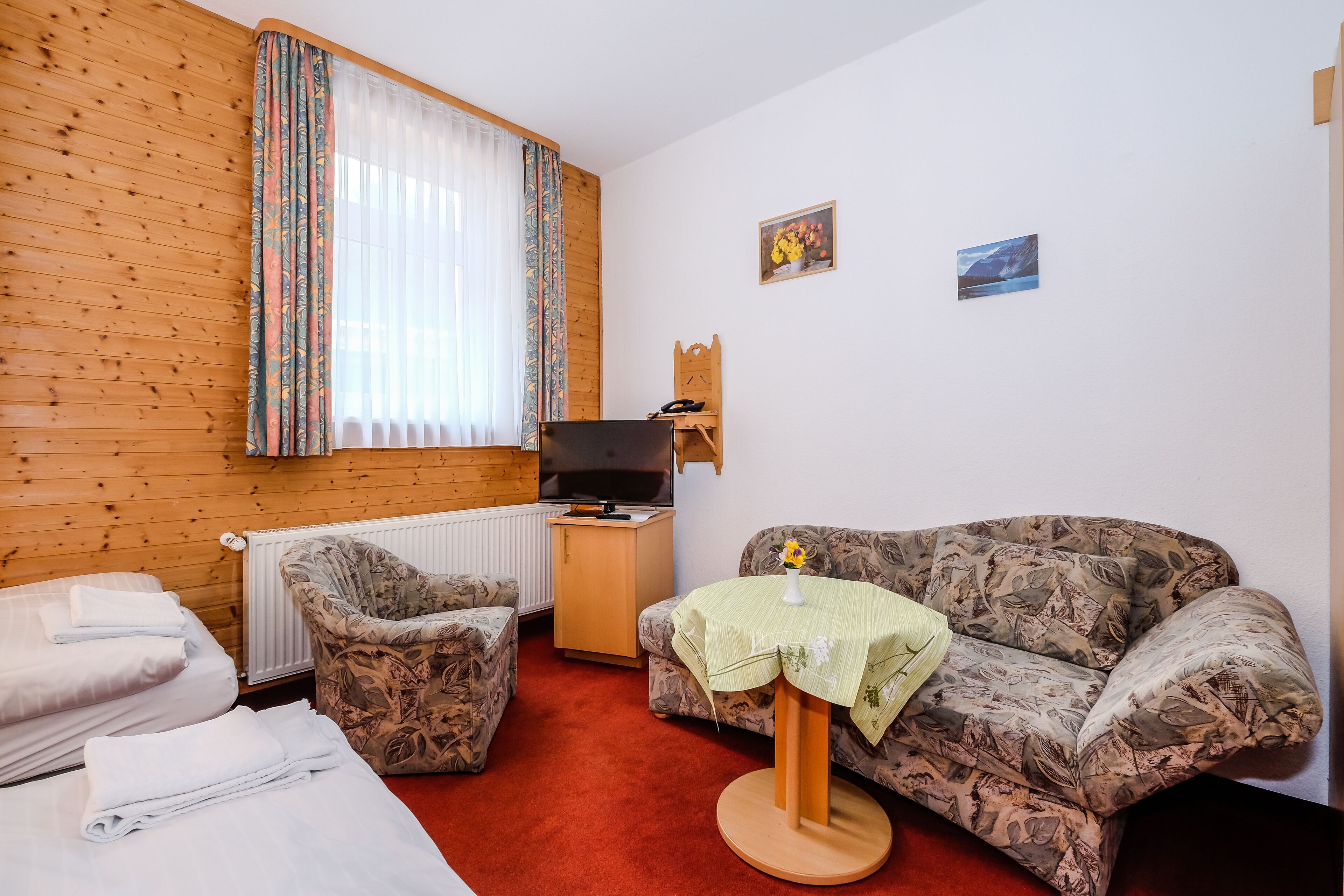 A room with a wooden wall, a sofa, a chair, a small table with a vase, a TV, and a radiator.