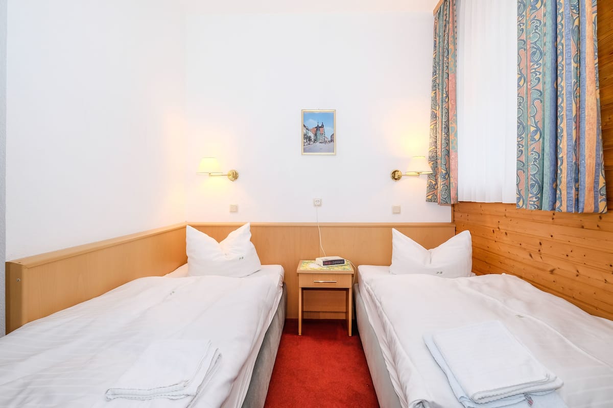 Habitación de hotel con dos camas individuales, cabecera de madera, mesita de noche pequeña con lámpara y un cuadro enmarcado en la pared.