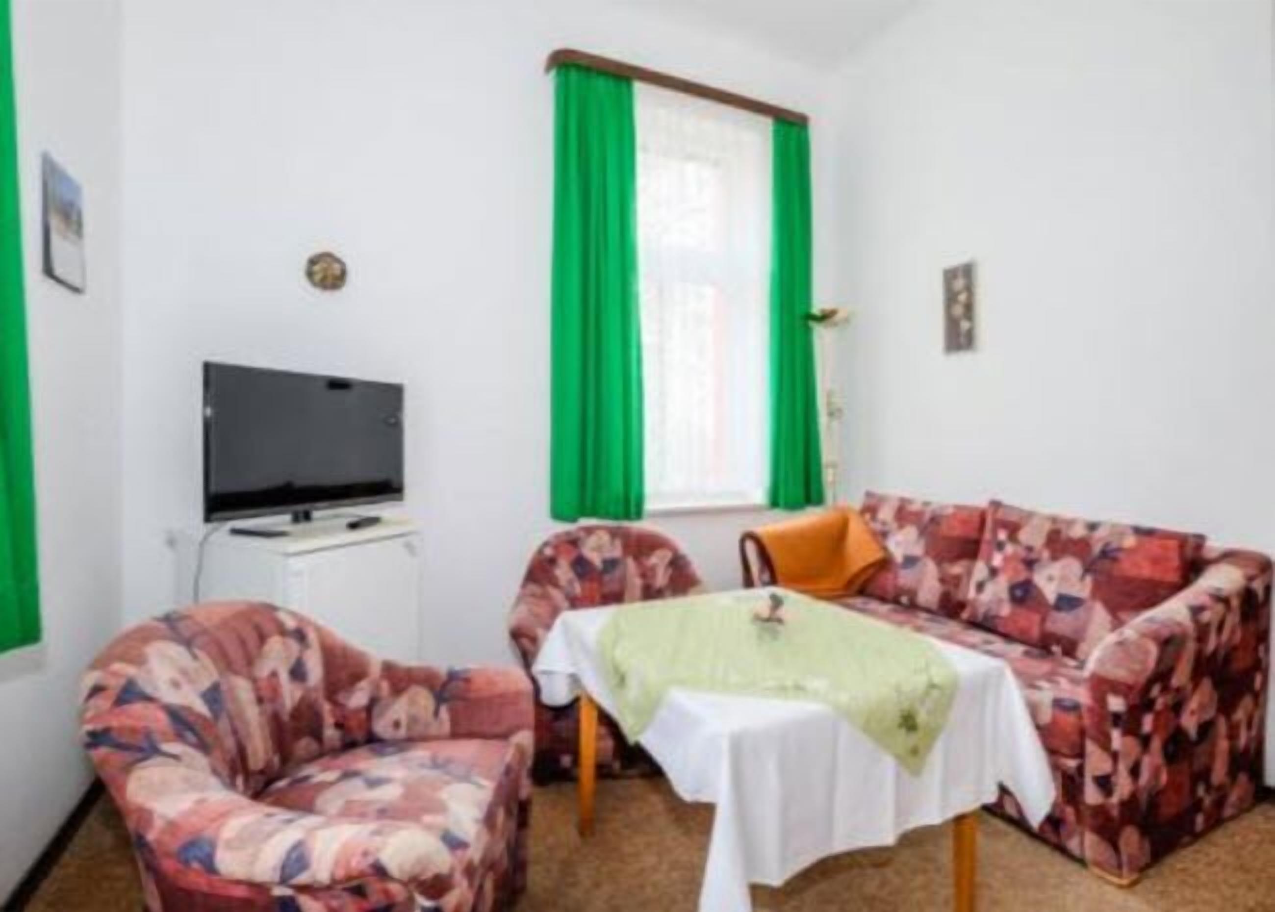 A small, cozy room with a sofa, two chairs, a table with a green tablecloth, a television on a stand, and a window with green curtains.