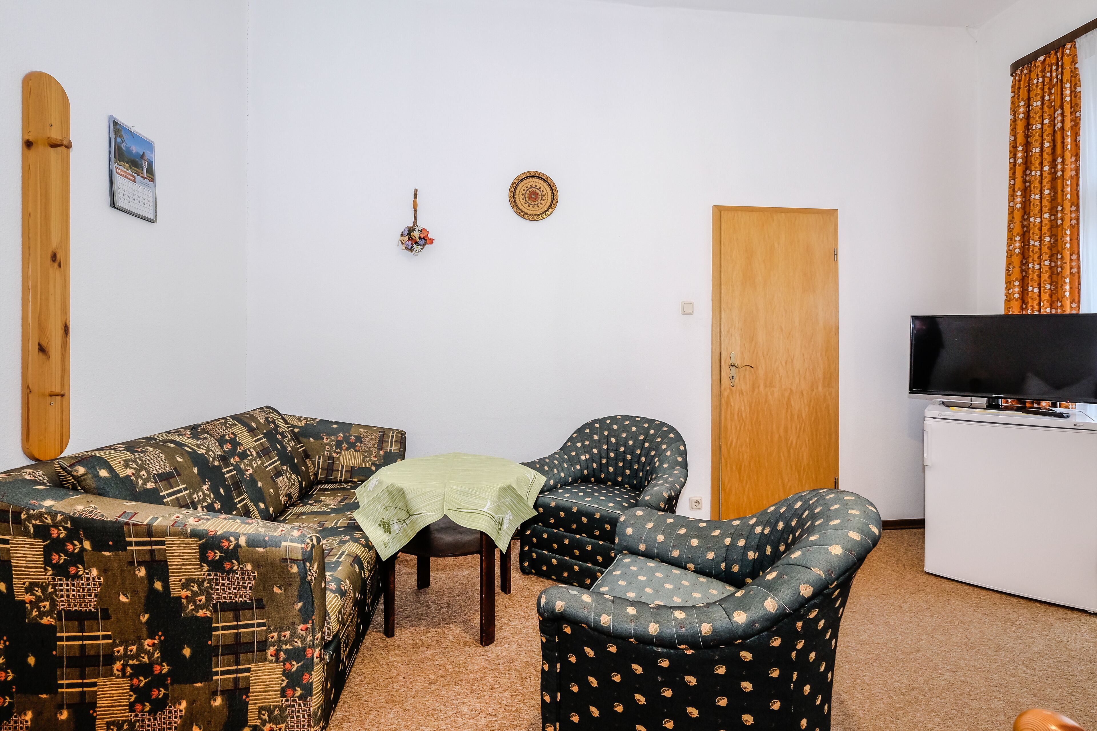 Ein Wohnzimmer mit einer Couch, zwei Sesseln, einem kleinen Tisch, einem Fernseher und einer Holztür.