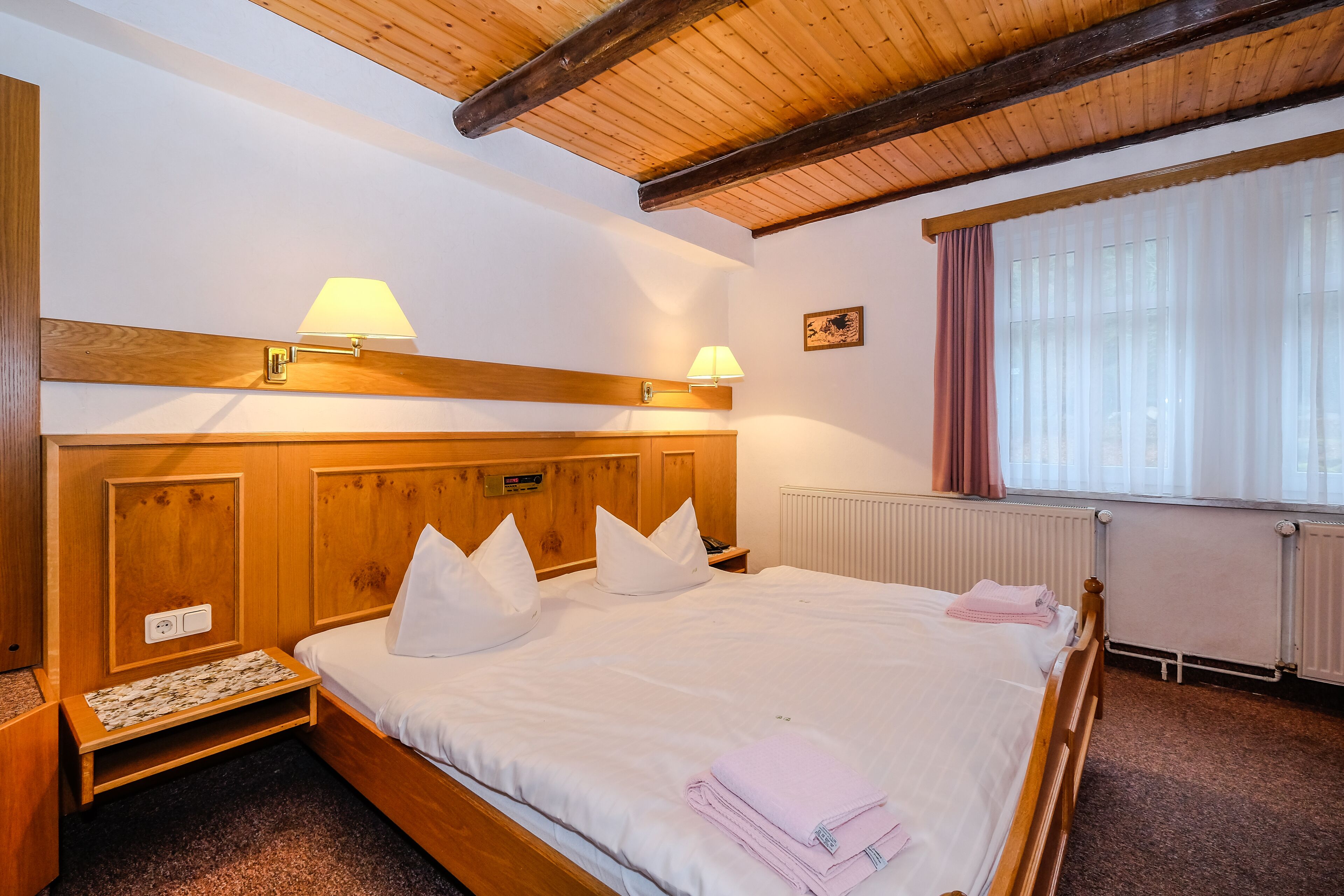 Una habitación de hotel con cabecera de madera, dos lámparas de noche, una cama con ropa blanca, una mesita de noche pequeña y una ventana con cortinas.