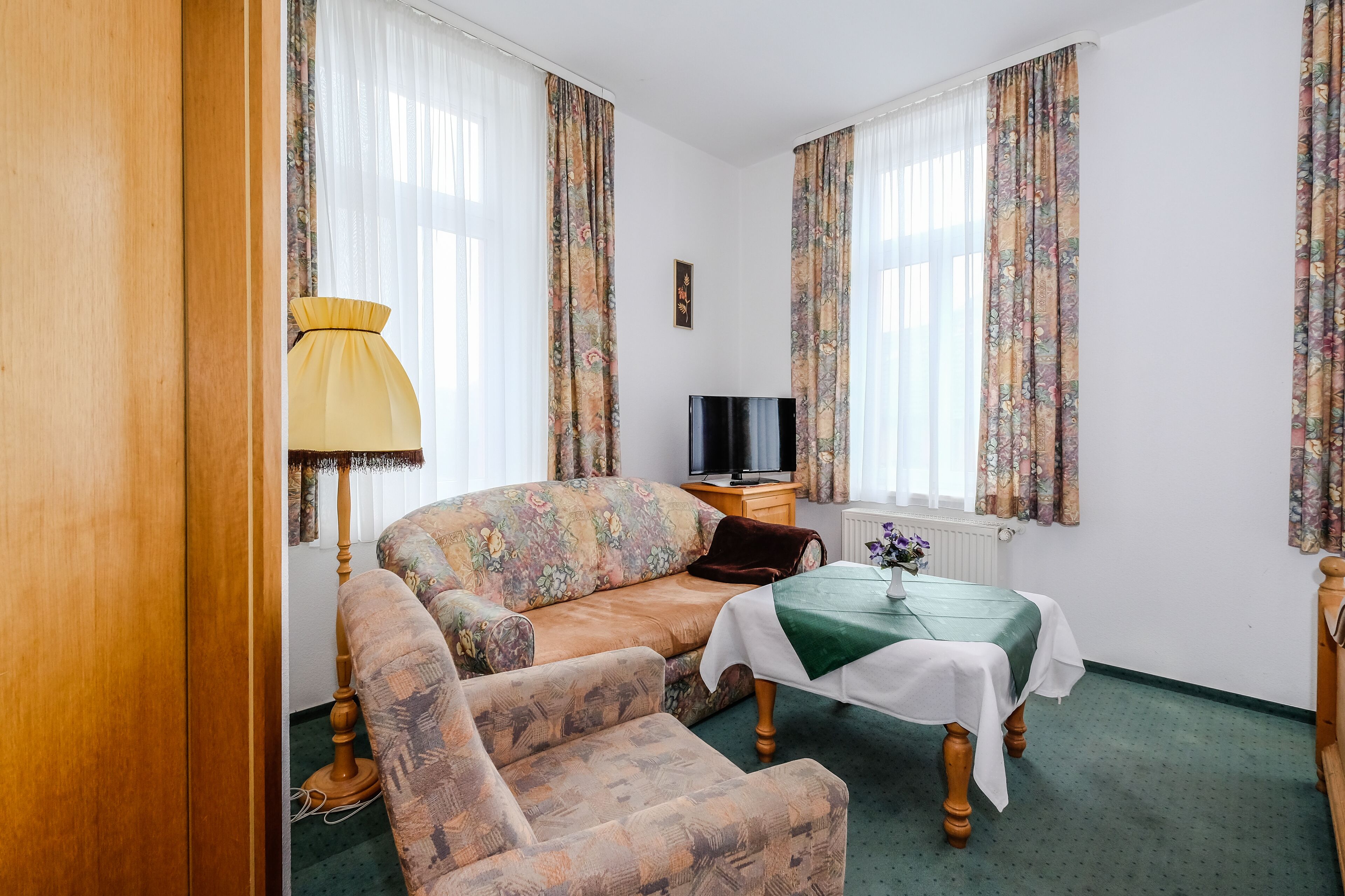 Una habitación con un sofá, una mesa pequeña con mantel verde, una lámpara y un televisor.